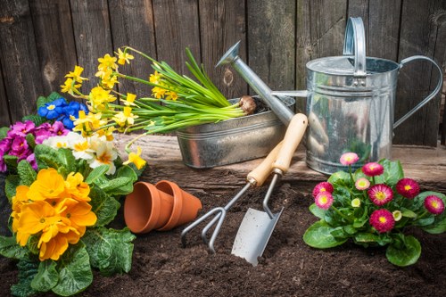The image depicts a neatly prepared garden bed with dark, moist soil ready for planting, situated in an outdoor garden area. Surrounding the bed are various gardening tools including a small hand rake, a trowel, and a pair of pruning shears, all positioned on the soil surface. To the left, there are vibrant flowering plants with yellow, purple, and pink blossoms, along with green foliage, adding colour and life to the scene. Behind the garden bed, a rustic wooden fence provides enclosure and privacy. A watering can is placed on the right side, suggesting ongoing garden maintenance, while a terracotta flowerpot lies on its side near the plants. In the background, there are additional plants and garden features, contributing to a well-maintained outdoor space typical of a home garden in Finchley. The scene appears to be set on a partly cloudy day, with natural light illuminating the garden elements, which aligns with professional gardening services offered by companies such as Garden Maintenance Finchley in the North London area.